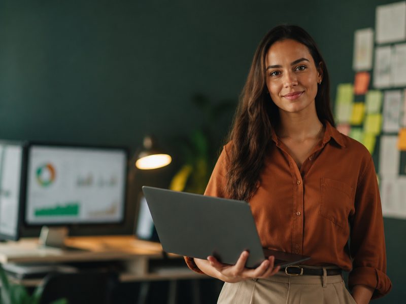 Profissional de marketing digital em escritório de agência, com um notebook aberto apoiado no antebraço